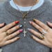 Hands with mismatched nail art in multiple colors and designs including orange, leopard print, red swirl, pink dots, light blue, and olive green almond nails styled with gold rings and a silver cross necklace.